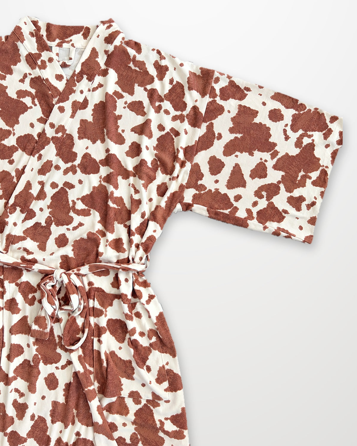 A long-sleeve wrap robe with a brown and cream cow print pattern, featuring a matching tie belt, laid flat on a light gray background.