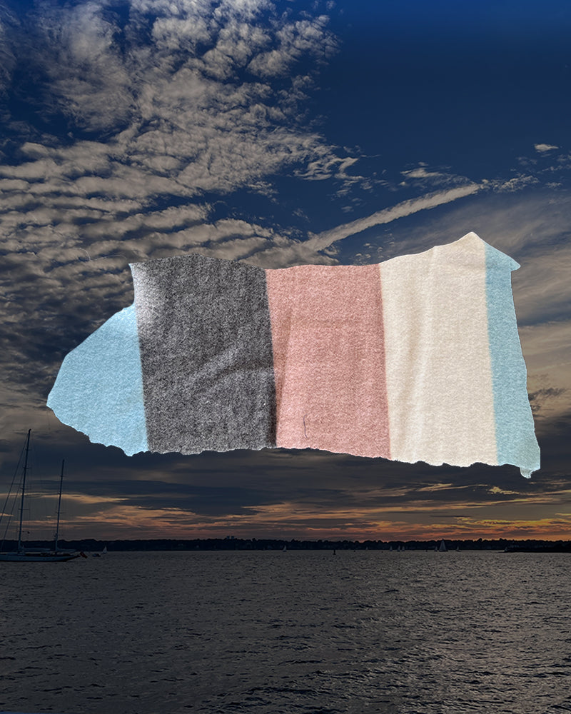 A fabric swatch sample with vertical color blocks in light blue, charcoal gray, dusty pink, and off-white, shown against a dramatic sunset sky over water.