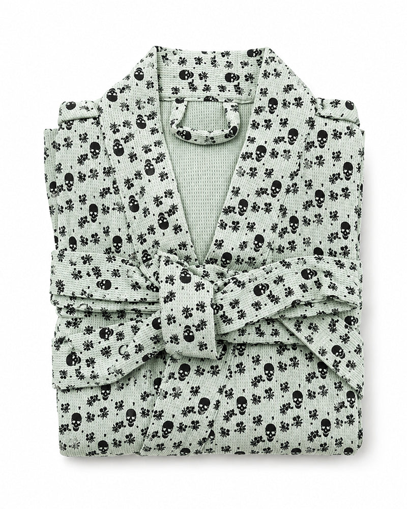 A folded light sage green robe with a black skull and crossbones pattern, featuring a matching tie belt and collar.